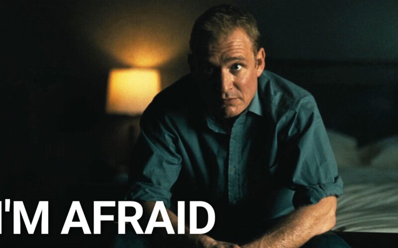 A man sits alone on the edge of a bed in a dim room at night, looking straight at the camera